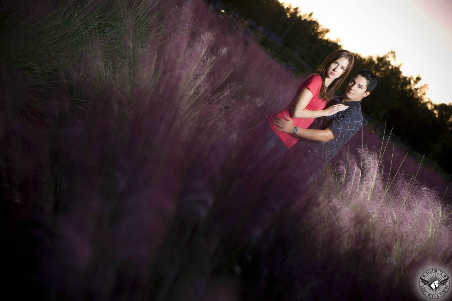 Ginger haired, blue eyed girl wearing a short sleeve scoop neck shirt and blue jeans is held by a Hispanic black haired guy with a blue grey shirt with a crosshatch pattern surrounded by the purple plumes of a reedy plant at dusk near the Long Center in this striking engagement photo in downtown Austin.